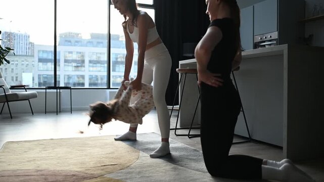 Mother and her friend helping a little girl with stretching exercises in a modern sunlit living room, promoting an active and healthy lifestyle from a young age through playful fitness