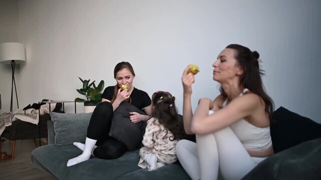 Candid moment of a females and little girl sitting on a sofa while chatting together and eating healthy pears in their cozy apartment living room