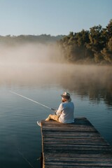 Fototapeta premium Older man fishing from wooden jetty at misty lake in morning