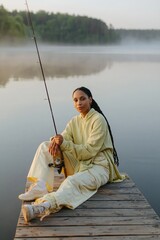 Fototapeta premium Woman sitting with fishing rod on misty lake dock at dawn