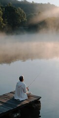 Fototapeta premium Person fishing sitting on pier at foggy lake sunrise
