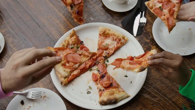 Group of anonymous young people sharing freshly baked pepperoni pizza reaching for slices at rustic table in cafe