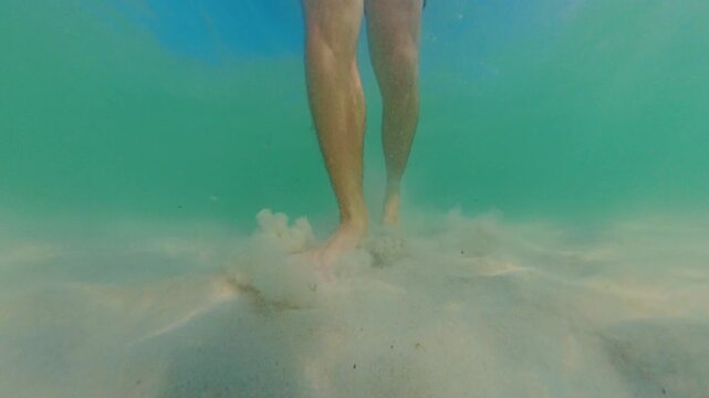 Close View Of Legs Walking Underwater, Foot Touching Sand And Lifting Sediment, Natural Light Filtering Through Water