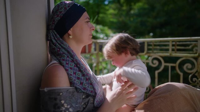 Mother in cancer treatment sits on balcony while restless toddler moves beside her, candid parenting moment showing fatigue, patience and emotional resilience in daily life