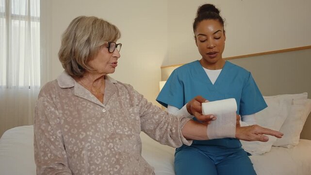 Attentive caregiver wrapping a gauze bandage around an elderly woman's injured wrist. Medical professional providing first aid and assistance to a senior patient recovering in her bedroom