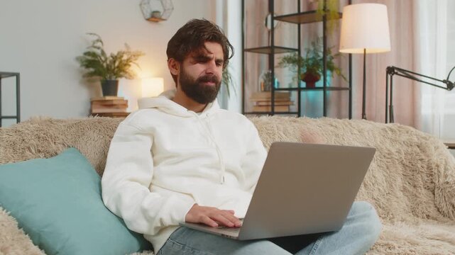 Upset young Indian young man sitting on sofa at home using laptop having anxiety and stress. Shocked bearded guy reacting to loss, failure bad news on netbook in living room apartment. Computer virus.