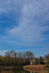 Obraz premium Airplane high in the sky above a calm lake, reeds and spring trees with dramatic clouds. 