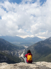 Fototapeta premium Female traveler sitting on cliff edge with yellow backpack overlooking mountain valley, dramatic clouds, snowy peaks, winding road create adventure travel scene with copy space, freedom concept