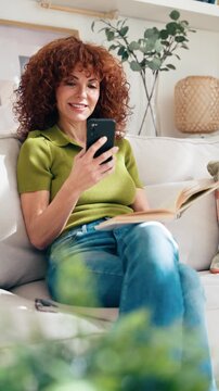 Video of mature smiling woman reading a book while using smartphone sitting on couch at home.