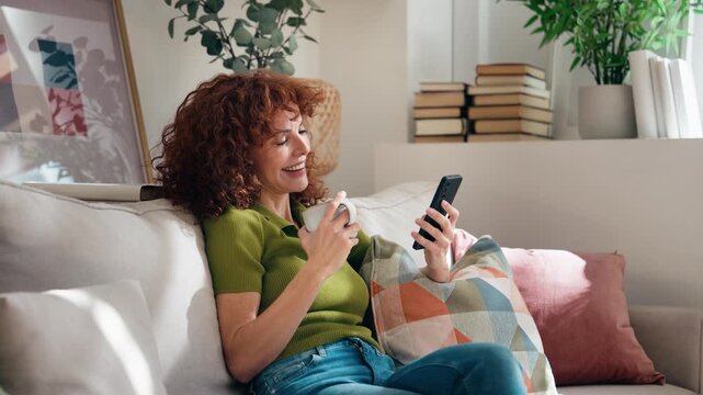 Video of mature pretty woman using smartphone while drinking a cup of coffee sitting on couch at home.