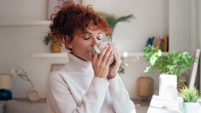 Video of beautiful mature woman drinking a cup of coffee while standing next to the window at home