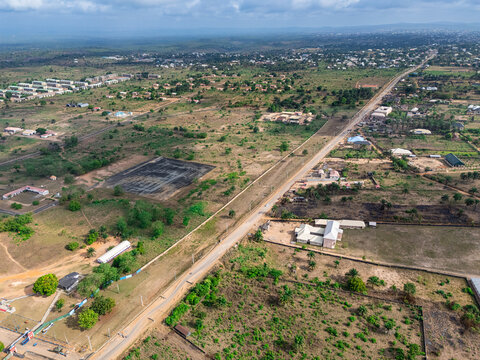 Aerial view of a straight road dissecting a vibrant green and brown landscape, featuring scattered buildings and a dark pond, Elu, Abia, Nigeria.