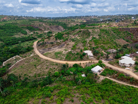 Aerial view of winding dirt roads cutting through verdant and cleared hillsides, dotted with a few light-roofed homes under a cloudy sky, Asaga, Abia, Nigeria.