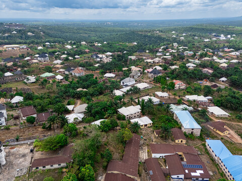 Aerial view of a sprawling settlement with diverse rooflines nestled amidst a verdant canopy, featuring a prominent blue-roofed building under a cloudy sky, Asaga, Abia, Nigeria.