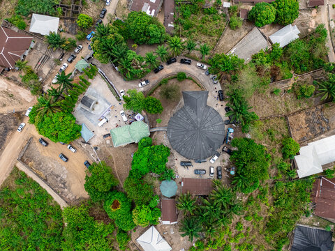 Aerial view of a circular church with a dark roof, amidst vibrant green foliage and dusty paths, bustling with parked cars, Asaga, Abia, Nigeria.