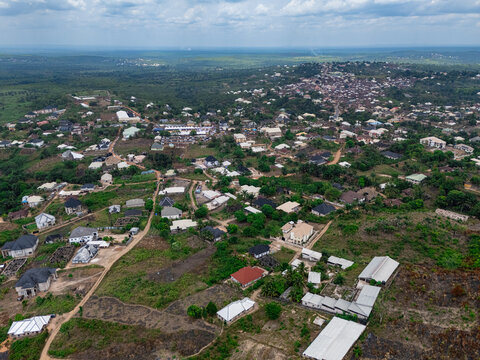 Aerial view of a sprawling settlement with diverse architecture nestled amidst verdant hills and earthy paths under hazy skies, Ohafia, Abia, Nigeria.