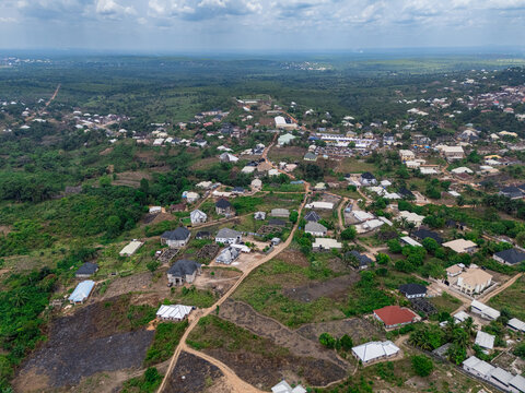 Aerial view of a residential area with scattered homes, verdant trees, reddish-brown cleared land under a cloudy sky, Ohafia, Abia, Nigeria.