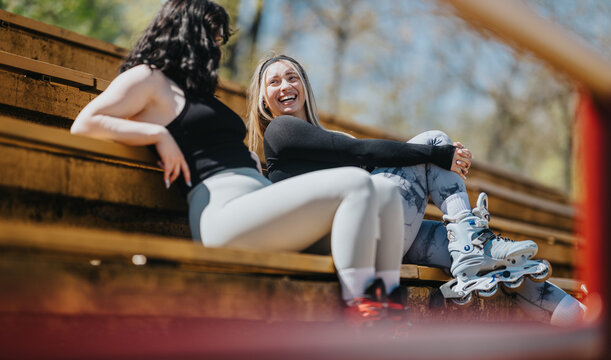 Two young women friends sit on outdoor bleachers wearing rollerblades and active wear, laughing together. They relax and chat on a sunny day after skating in the park.