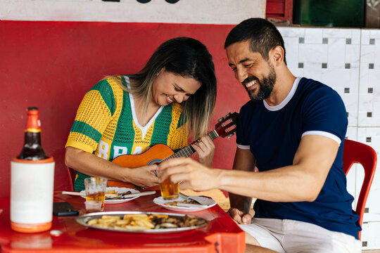 Brazilian couple enjoying time at a casual bar, smiling while playing cavaquinho and sharing drinks and food in a relaxed and vibrant social atmosphere.
