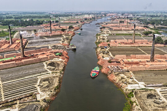 Aerial view of a stark industrial landscape where countless red brick kilns belch smoke into the hazy sky, flanking the dark Buriganga River, Narayanganj, Dhaka Division, Bangladesh.