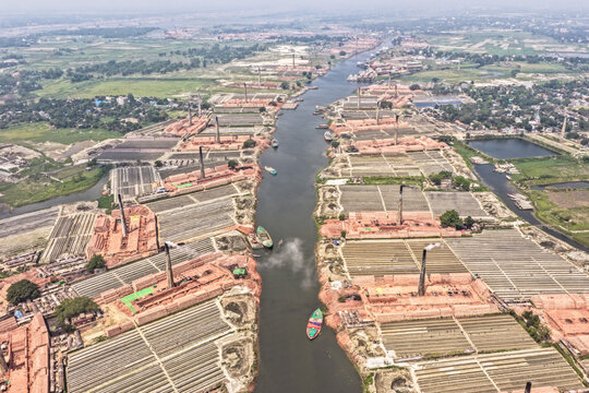 Aerial view of the dark Buriganga River winding through a dense tapestry of reddish-brown brick kilns, chimneys spewing smoke into the hazy air, Narayanganj, Dhaka Division, Bangladesh.