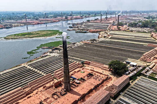Aerial view of the Buriganga River, its banks a striking expanse of red brick kilns with smoke plumes rising, Narayanganj, Dhaka Division, Bangladesh.