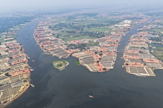 Aerial view of countless reddish-brown brick kilns lining the dark, winding Buriganga River, with a lone green island amidst the industrial landscape, Narayanganj, Dhaka Division, Bangladesh.