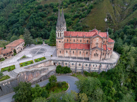 Aerial view of the Basilica de Santa Maria la Real de Covadonga, its red tiled roof contrasting with the green forest, stands majestically atop a stone precipice, Covadonga, Spain.