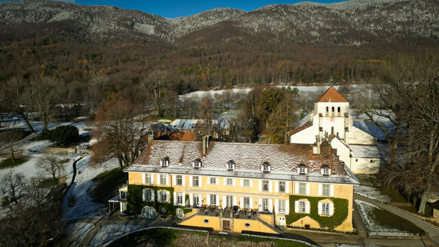 Aerial view of the grand, mustard-yellow building adorned with climbing ivy, standing in contrast to the snow-dusted ground and the towering mountains in the background, Cheserex, Vaud, Switzerland.
