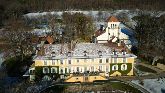 Aerial view of a grand, snow-dusted building with a tower, framed by winter trees and patches of snow-covered ground, Cheserex, Vaud, Switzerland.