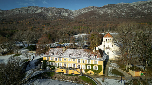 Aerial view of a stately yellow building with a red-tiled roof, contrasted against the snow-dusted landscape and the backdrop of the mountains, Cheserex, Vaud, Switzerland.