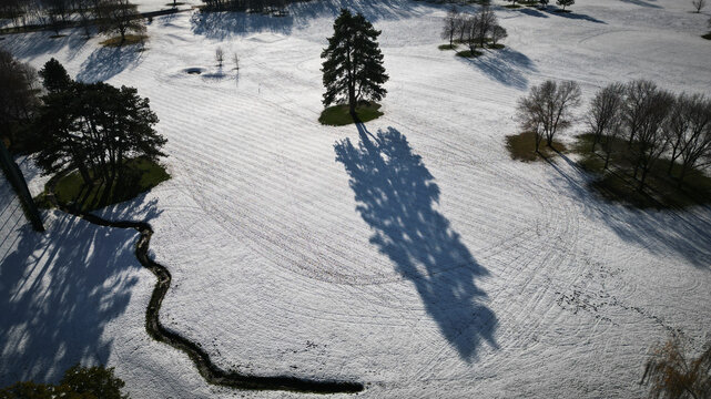 Aerial view of a snow-covered landscape with trees casting long shadows, and a winding stream cutting through the white expanse, Cheserex, Vaud, Switzerland.