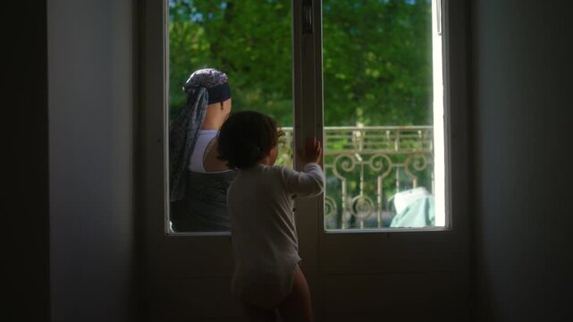Toddler taps on glass window seeking mother attention while she turns and gestures for quiet, candid parenting moment showing communication, boundaries and playful interaction
