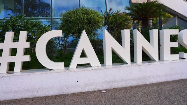 Daytime pan reveals oversized white 3D hashtag CANNES letters on a concrete plinth at Palais des Festivals, with palm trees, shrubs, and reflective glass highlights.