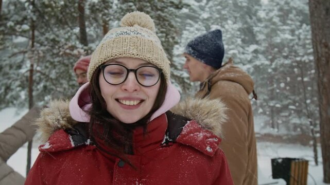 Chest-up portrait of millennial girl wearing bobble hat, glasses and red puffer jacket posing for camera on winter day, with friends in background