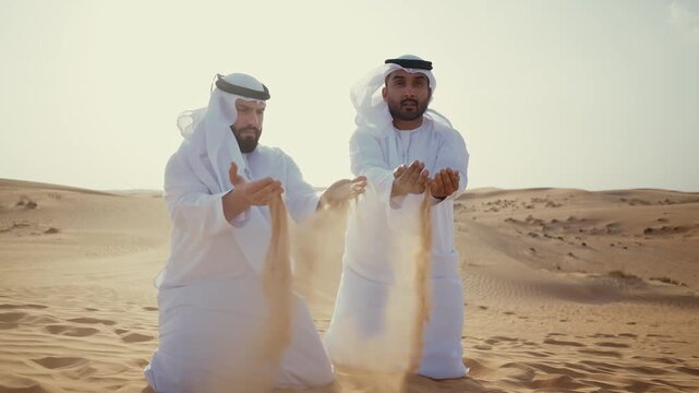 emirati men kneeling in the desert, wearing traditional white kandura, letting golden sand run through his open hands while enjoying the beautiful arid landscape and bright sunshine