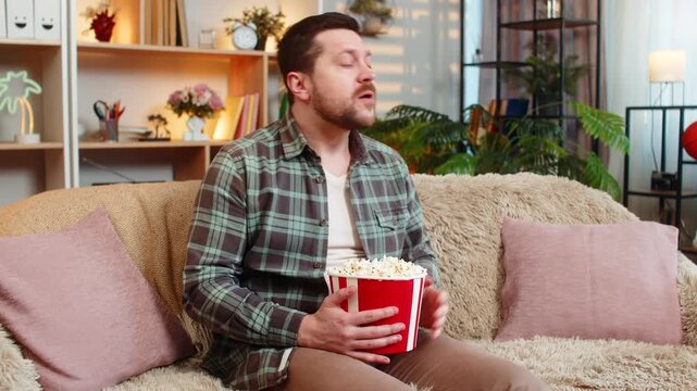 Young man on home sofa watches sports with popcorn and cheers with raised fist after big winning moment indoors. Adult guy on home sofa reacts with joy to victory and enjoys match night entertainment.