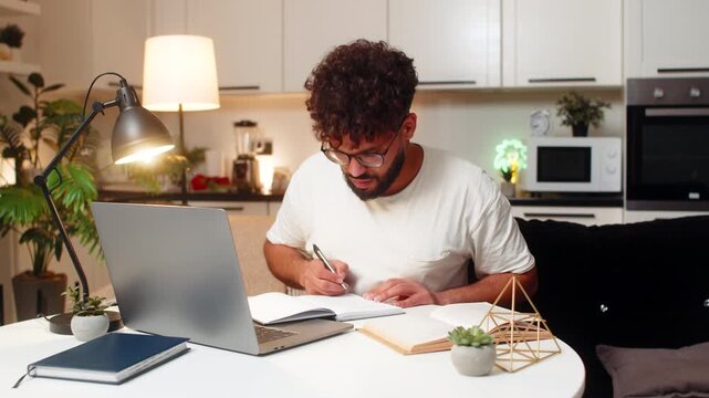 Arabian man at home studies online on laptop watches lesson writes notes smiles learns for exam. Egyptian guy sitting at table reads book solves test in notebook prepares revision feels tonight calm.