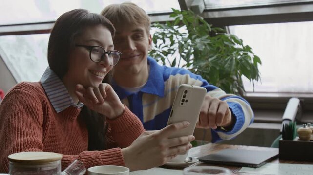 Millennial girl with glasses and gen Z male friend looking at smartphone with beige case while visiting restaurant