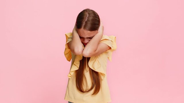 Young child girl covers ears with both hands, opening eyes wide in sudden fear panic caused by a loud sound or noise. School kid isolated on pink background, reacting emotionally with dramatic gesture