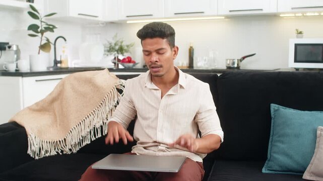 Indian man at home saves files completes backup closes laptop and shows thumbs up with relief. Arabian guy on sofa leans back smiles at camera breathes slowly and ends day with final wrap up and rest