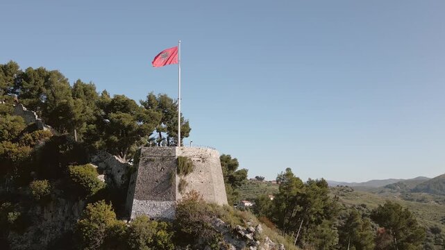 Aerial view Albanian national flag prominently wave atop stone fortification at Berat Castle - Kalaja e Beratit under clear blue sky, overlooking lush, forested hillside and Berat cityscape