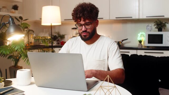 Arabian man at home opens laptop starts freelance project types checks deadline remote work. Egyptian guy sitting at table uses trackpad edits files answers email manages tasks drinks tea calm night