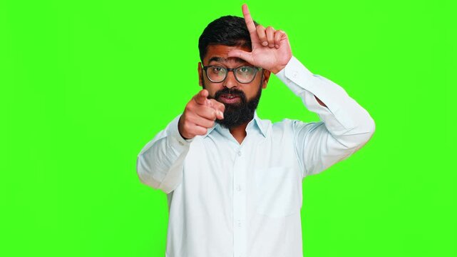 Indian man makes an L sign on his forehead with fingers while smiling playfully, teasing viewer as loser. Arabian guy isolated on green chroma key background showing joke, sarcasm and mock gesture.