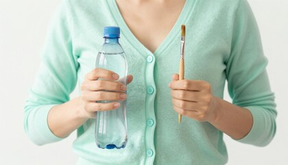 Fototapeta premium Person wearing a mint cardigan holds a plastic water bottle in one hand and a paintbrush in the other, photographed as a close cropped torso shot