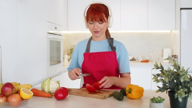 Young woman at home wears headphones plays anti stress music and cuts tomatoes for salad on board. Redhead girl in kitchen keeps rhythm uses knife and bowl calm breath and neat slices and gentle mood.