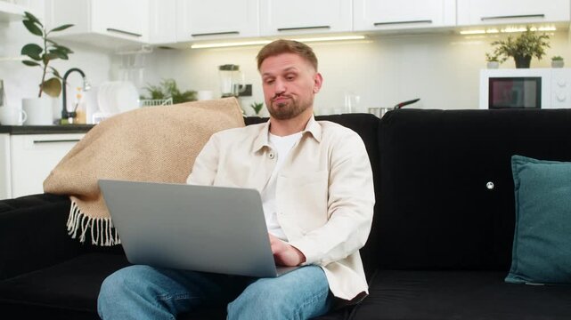 Young man at home works on laptop feels tired and demotivated and takes deep sigh during hard task. Caucasian guy on sofa slumps shoulders shows fatigue stress low energy and tries to focus now again