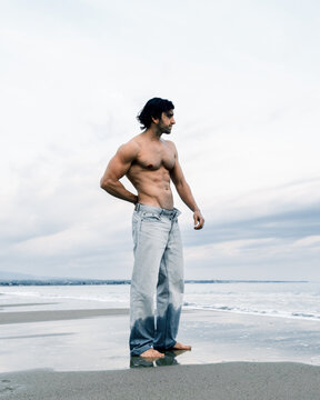 Muscular man in wet jeans standing on a beach with the ocean in the background