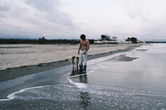 Man and his dog walk along the wet sand at the edge of the sea on a cloudy day