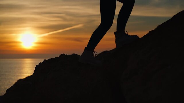 Silhouette of female hiker show legs and boot as climbs rocky mountain path with trekking poles, against stunning sunset over calm sea and peaceful natural horizon